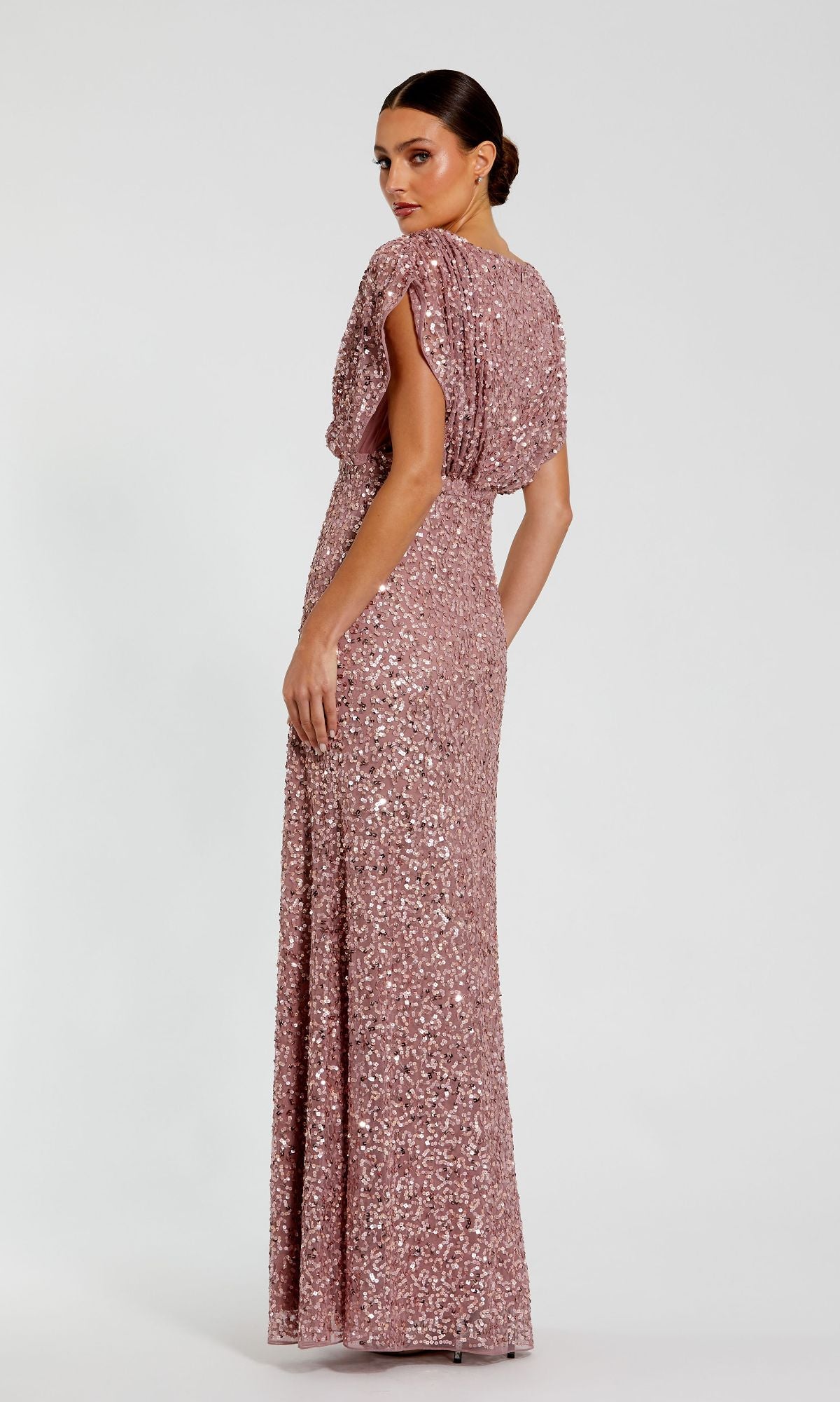 A woman faces away, wearing the Mac Duggal 5640 Short Sleeve Long Sequin Formal Dress in rose gold; this floor-length gown features short sleeves and a fitted silhouette, set against a plain white background.